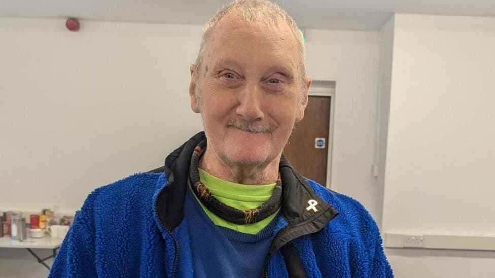 An older person wearing a blue fleece jacket over several layers of clothing stands indoors in a community facility. Tables and shelves with food and household items can be seen in the background.