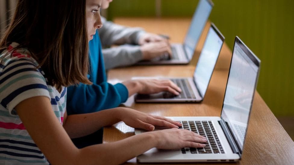 A girl with shoulder length dark hair and a stripy T-shirt sits next to two other children at a table with laptops.