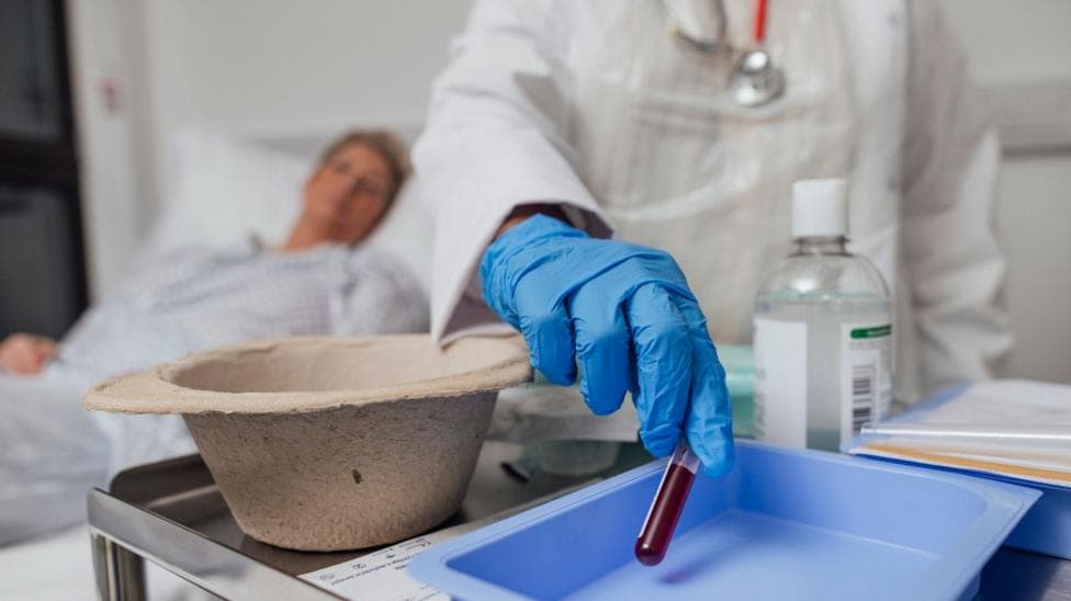 A close up of a nurse's hand taking a blood sample from a woman who is undergoing treatment for cancer. The nurse is wearing a blue plastic glove and holding a test tube half filled with blood.