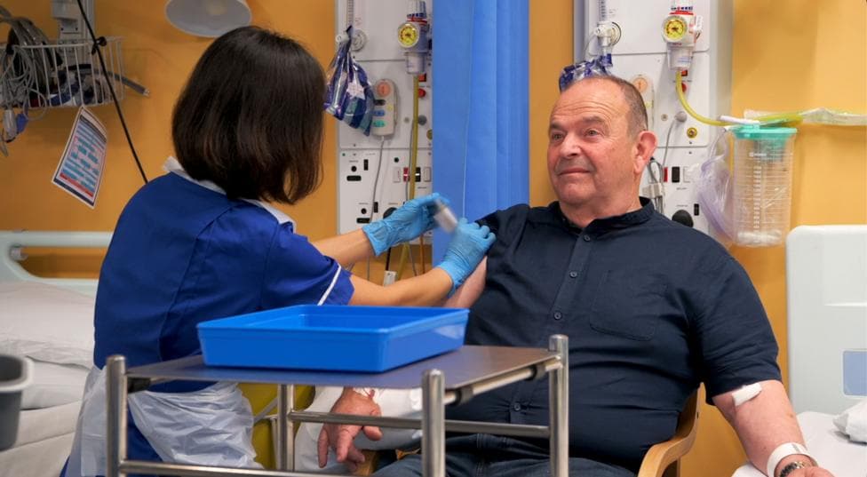 Tony Chapman sat in a hospital chair being given an injection. He is wearing a dark blue shirt. A a nurse in a blue uniform is giving him an injection of either the vaccine treatment or a placebo drug.