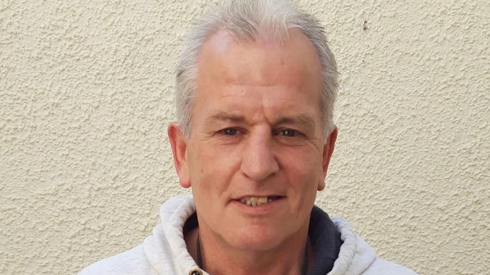 Clifford Foster stands in front of a white wall. He wears a white hoodie, has silver hair and brown eyes and smiles at the camera.