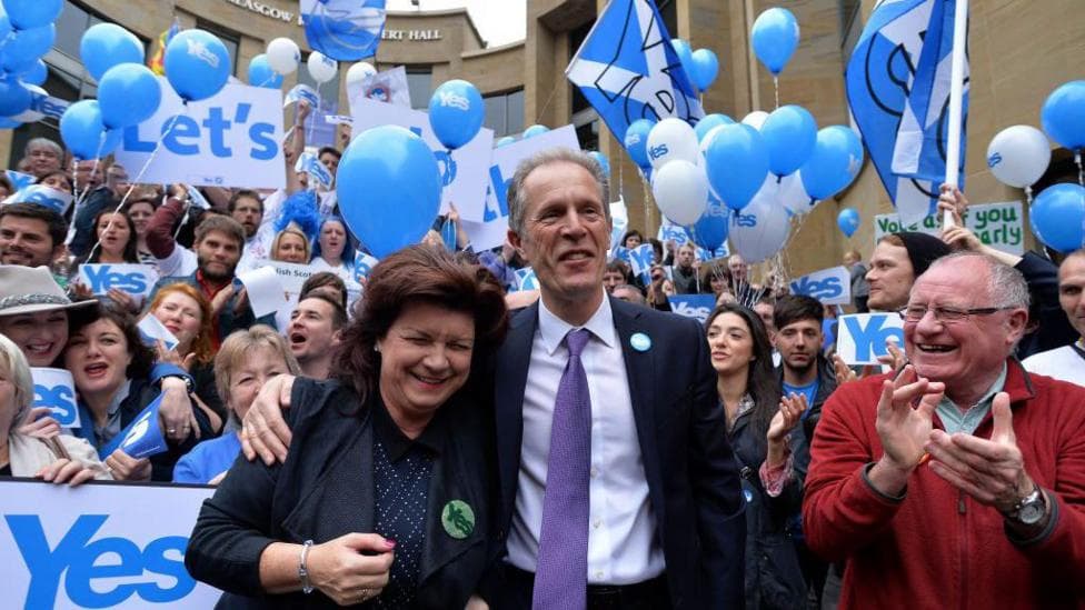 Blair Jenkins, is wearing a dark suit, white shirt and purple tie. He is smiling and has his arm around actress Elaine C Smith who is laughing. Dennis Canavan stands to his right, and is smiling and clapping. They are standing in front of a cheering crowd of people with Yes campaign banners