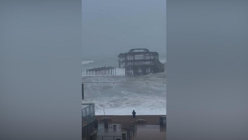 Part of a pier, which is black and a skeleton structure, falls into a rough and windy sea.