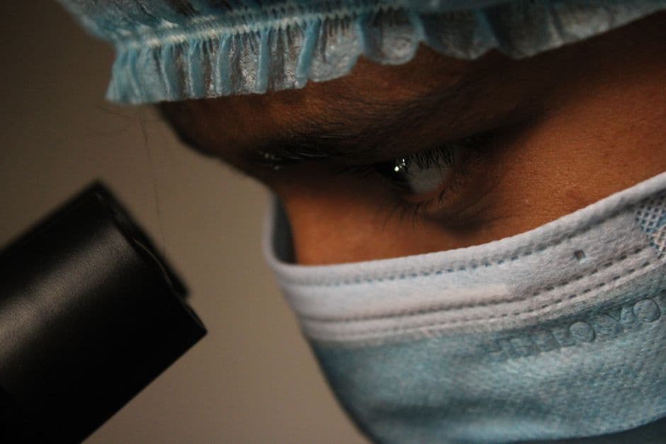 Close-up of a scientist wearing a mask and cap observing through a microscope.