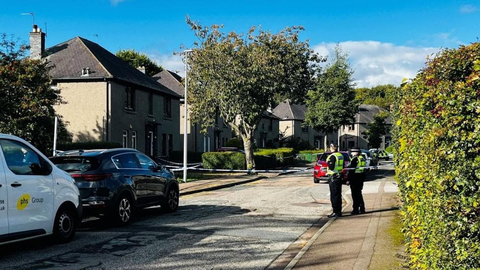 Police officers and police tape in an Aberdeen street, houses and hedges can be seen.
