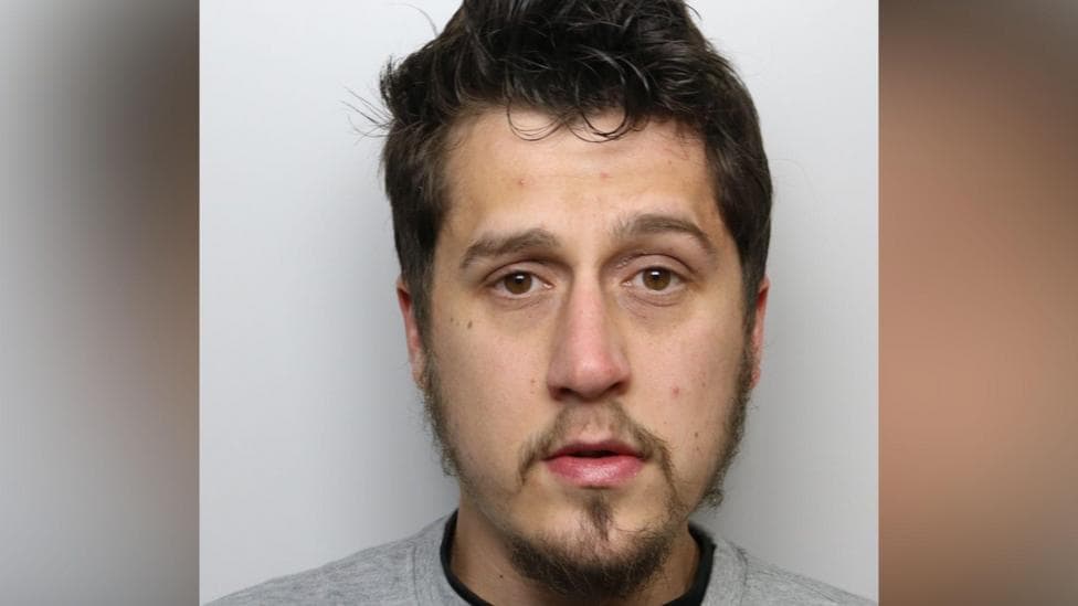 A man with dark hair and a goatee beard wearing a police-issue grey sweatshirt.