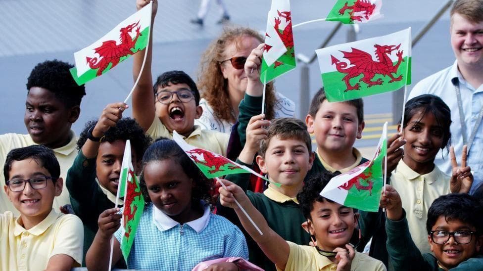 A group of children of different ethnicity waving small Welsh flags. They are smiling and cheering while looking at the camera.