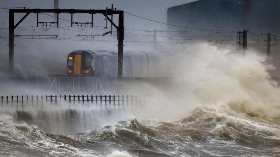 A train travels along a coastal railway as huge waves crash over stormy seas, spraying water across the tracks.