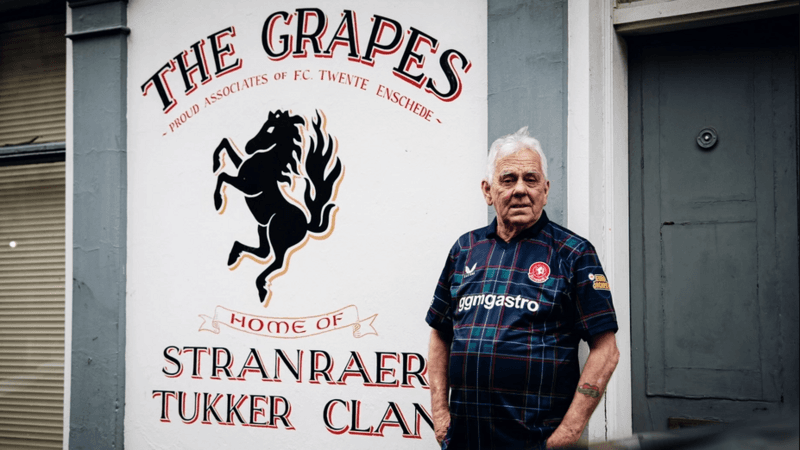 Billy Hodge standing outside The Grapes pub in Stranraer. It has the name of the bar in black text on a white background behind him and a large icon of a horse. He is wearing the shirt and looking slightly off camera.