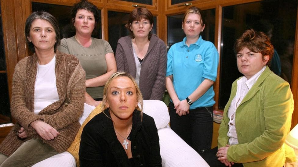Six women stare at the camera. The one on the far left has short black hair, wearing a white T-shirt and brown cardigan. The woman beside her has short black hair and is wearing a green
T-shirt. The woman in the middle has short brown hair and is wearing a brown cardigan. The woman second from right is wearing a blue T-shirt and has dark hair which is tied up. The woman on the far right sits on a stool. She is wearing a white blouse, green jacket and has short, dark hair. The woman at the very front is sitting on a large white armchair. She has long blonde hair and is wearing a dark top with a large silver cross and chain around her neck.