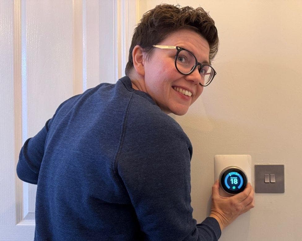 Cora Harrison, a woman with short brown hair and glasses in a navy jumper, adjusts the temperature on a thermostat to 18 degrees