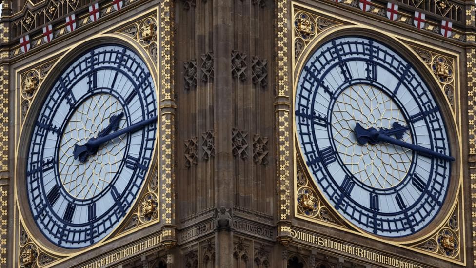 A close up of two of the clock faces on Big Ben. It is a large, ornamental clock with gold and black detailing on the top of the brown Elizabeth Tower. The black clock hands are showing a time of quarter past two. Along the top of the clock face are small red and white St George's Cross flags.