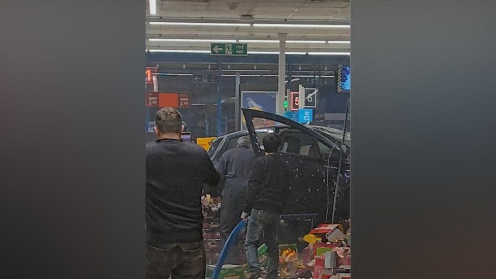 A car inside a supermarket with the backs of three people shown stood near it.
