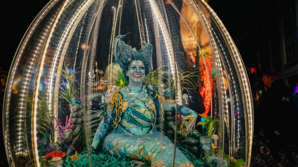 A performer in an elaborate blue-green costume resembling a mythical underwater creature is displayed inside a transparent, light-filled dome during the Paisley Halloween Festival. The float is decorated with plants, coral-like shapes and sea-themed ornaments, while bright white lights outline the dome structure.