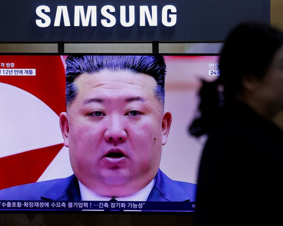 A silhouetted figure passes a TV screen showing Kim Jong Un speaking, below a Samsung sign