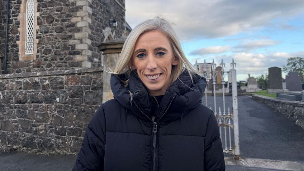 Woman with blonde hair wearing a dark coat standing in front of a church. There is a stone wall to her left, a silver gate behind her and a graveyard is visible behind her.