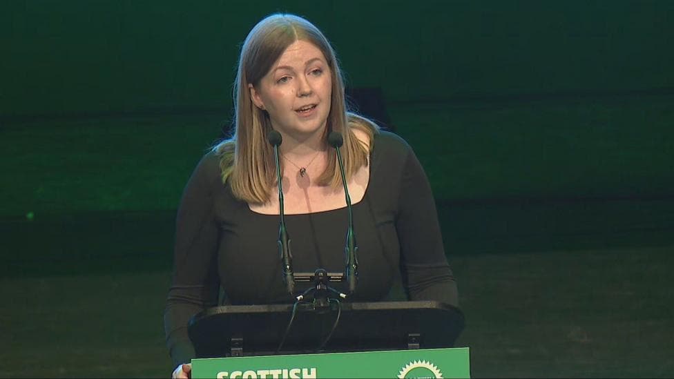 Gillian Mackay stands at a podium with Scottish Greens branding, addressing delegates on a conference stage illuminated with green lighting.