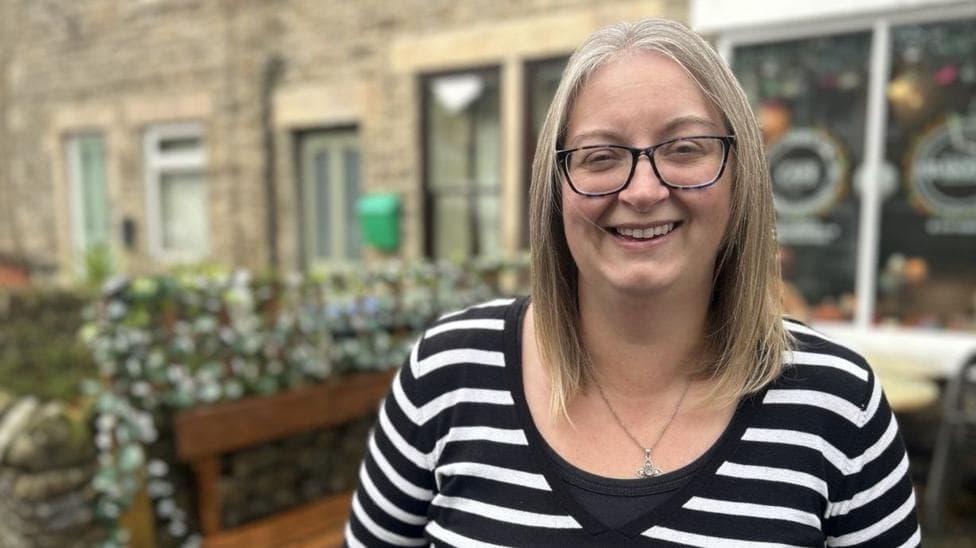 A woman with a black and white stripy top on is standing on the high street, she is wearing black rimmed glasses and has a silver necklace on. Behind her is a cafe and a row of stone cottages. She has fair shoulder length hair in a middle parting.