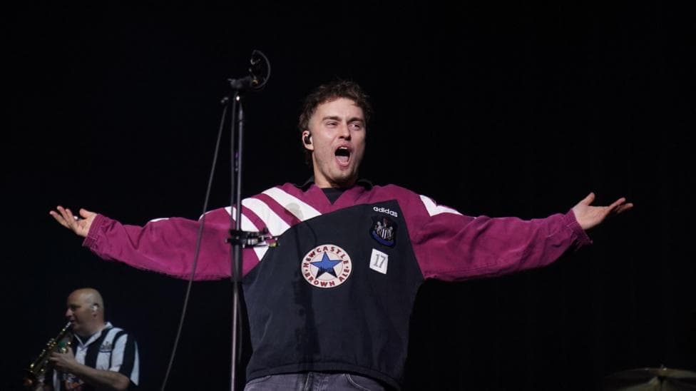 Sam Fender on stage. He is wearing a Newcastle United top which contains the club badge as well as the Newcastle Brown Ale star. A saxophone player in the background is wearing a Newcastle United shirt.