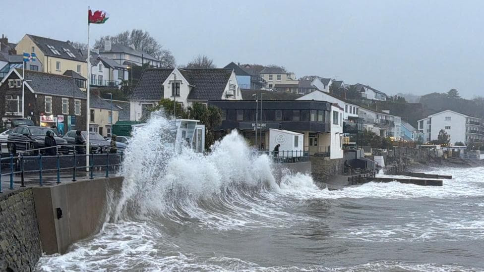 Large waves splash on to the sea wall and the promenade, with several people wearing coats looking on from behind another wall further back. Houses and buildings and a Welsh flag can be seen.
