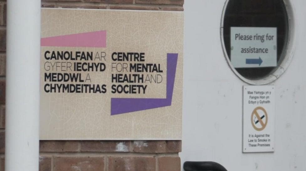 Close up of the entrance to the Centre for Mental Health and Society office in Wrexham. A bilingual sign is attached to a brick wall, to the left of a white door with a porthole window, which is mainly filled by a sign saying "please ring for assistance." The main body of the door has a bilingual no smoking sign on it.