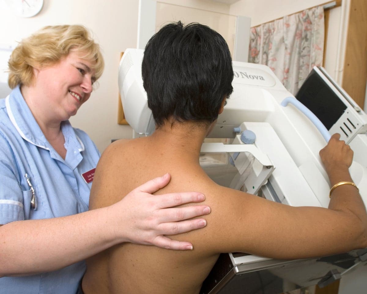 A nurse smiles with her hand on the shoulder of an Asian woman undergoing a mammogram