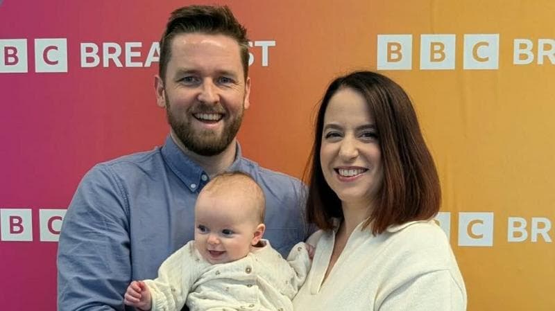 Kate and Matt are holding their baby daughter and are smiling at the camera in front of a BBC Breakfast-branded sign.