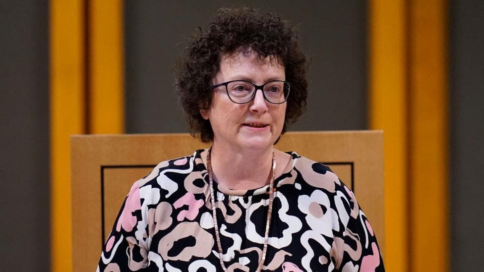 Elin Jones wears a possibly floral dress which is black, white, beige and pink.
She has a long necklace, perhaps of a precious metal, black spectacles and curly hair