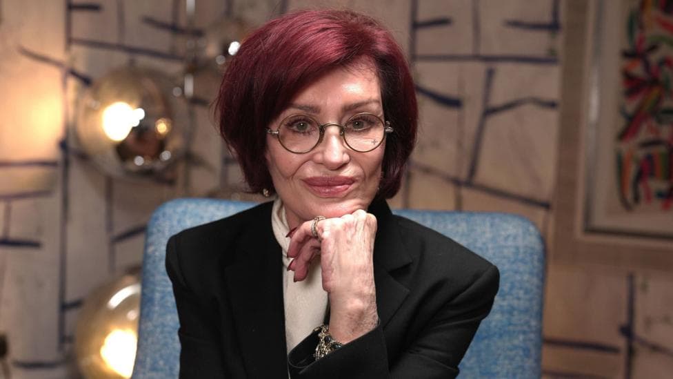 Sharon Osbourne poses with her chin resting on her hand, against a colourful background