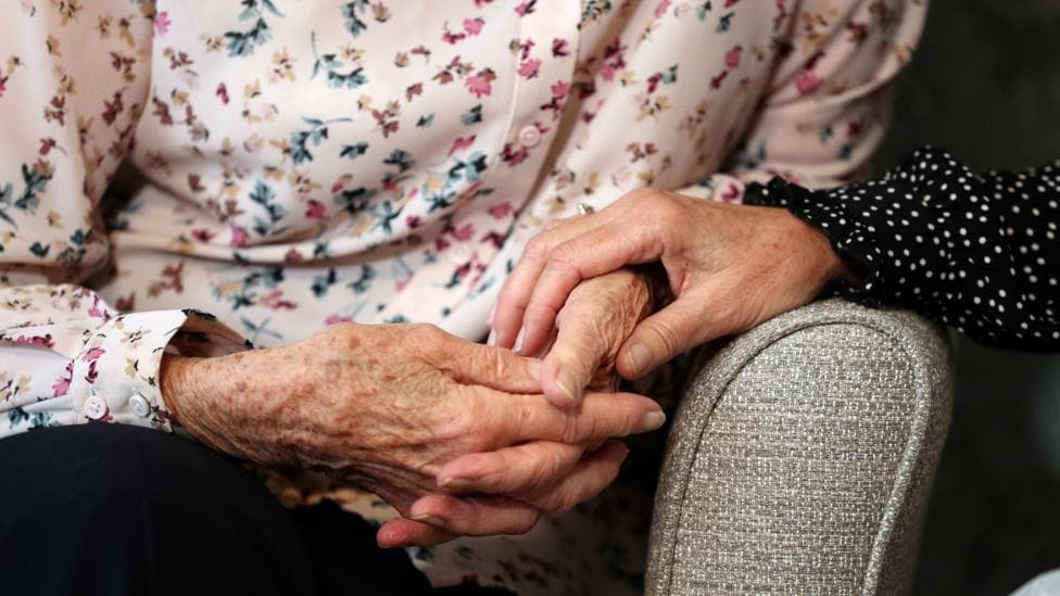A woman sits in a chair in a care home, while another woman holds her hand.