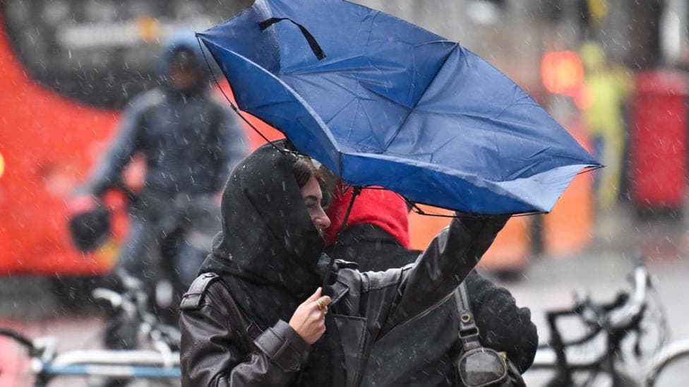 A woman in a raincoat struggles with an umbrella that has turned inside out, with a blurred city street scene with buses and bicycles in the background and rain falling overhead.