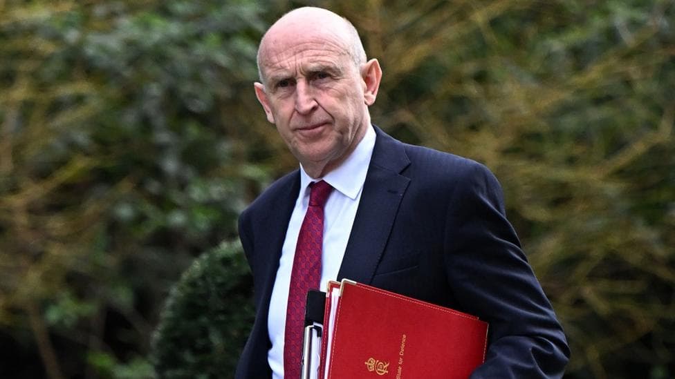 John Healey walks along Downing Street with files under his arm and wearing a dark suit and a red patterned tie.