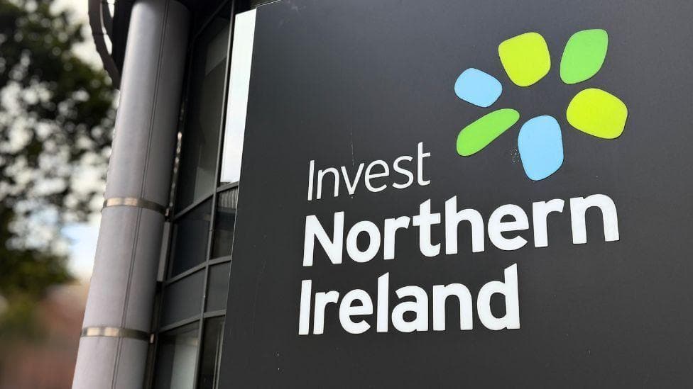 Close shot of Invest Northern Ireland logo on a grey board. It has a blue and green spiral logo. Exterior of glass building and out of focus tree in the background
