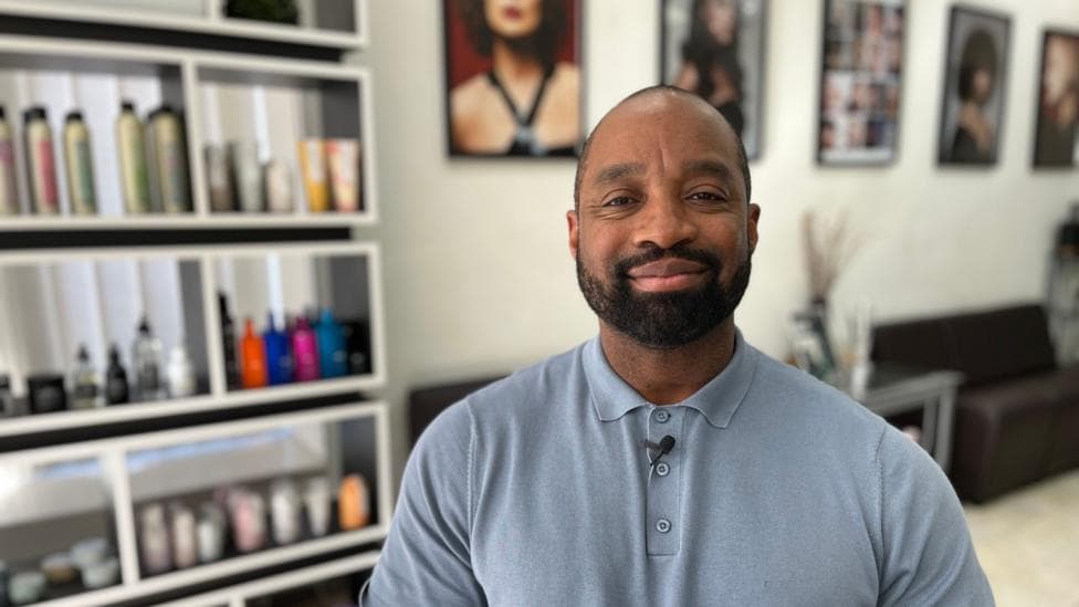 Marcus is sitting on a chair in his hair and beauty salon. He is black, with short hair and a moustache and beard. He is wearing a blue polo shirt. Behind him are shelves full of products and prints of hair models on the wall.