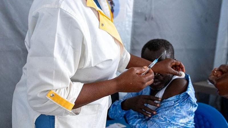 A health worker in a white lab coat holds a syringe as she prepares to administer a dose on a man's shoulder,
