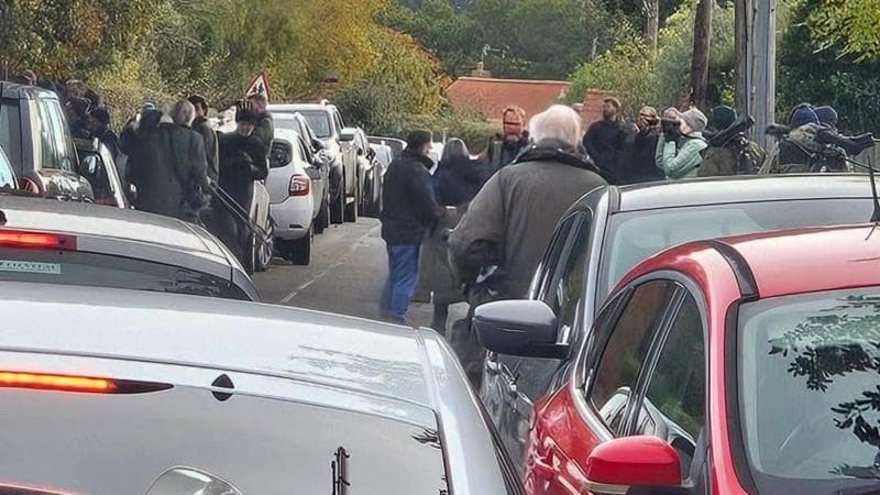 Traffic congestion with cars parked on either side of a road and others in the middle of the road. There are people with binoculars, telescopes and tripods standing on the pavements and in the road.