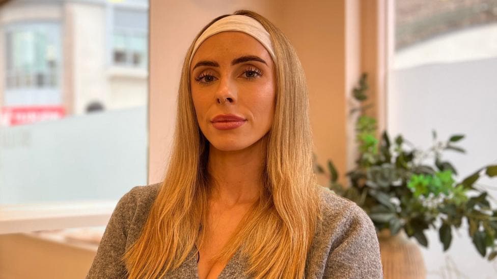 Meg Hamon is seated in a salon or clinic chair, wearing a grey wrap-style top and a light-coloured headband. A large wall mirror reflects part of the room, while windows and a potted plant are visible in the softly lit background.