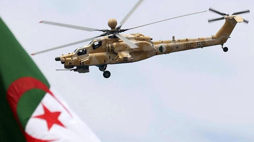 A military helicopter painted in beige camouflage with white patches flies above an Algerian flag, which has green and white halves and a red crescent and star in the centre.