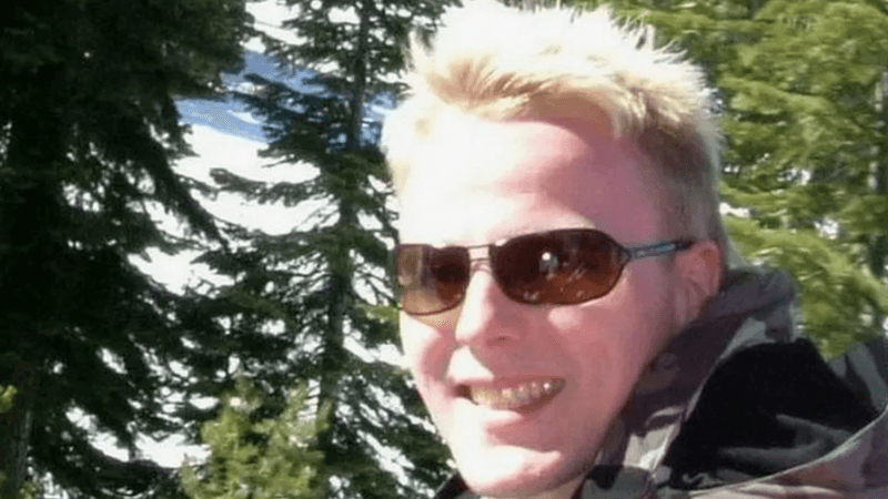 A man with blond hair and wearing sun glasses is smiling at the camera with green trees and snowy hills in the background