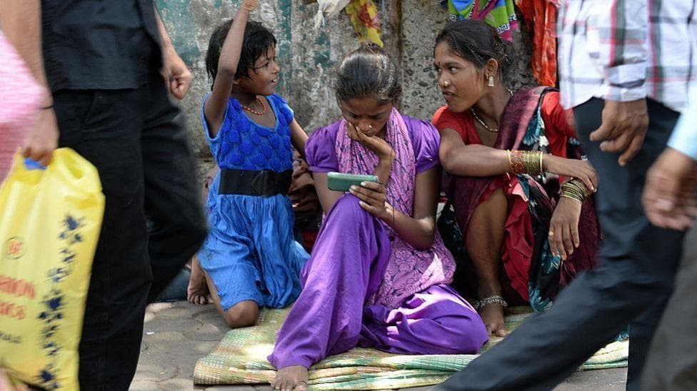 A young female Indian pavement dweller watches a movie on a mobile phone with two other women besides her.