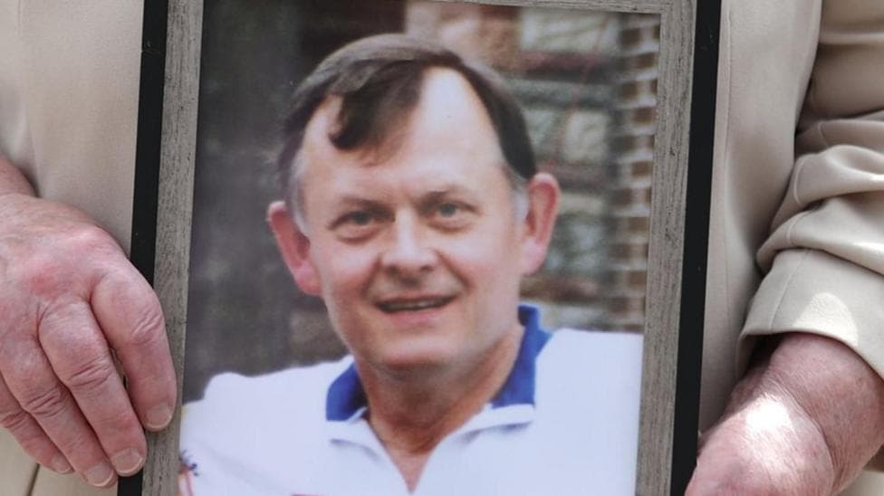A photo of Sean Brown being held, he is a man with dark hair, wearing a white top with blue trim.