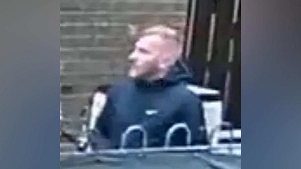 A grainy CCTV image of a fair haired man wearing a black jacket.