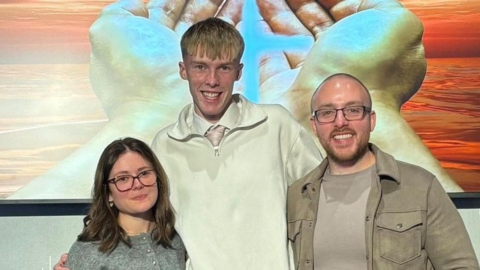 Three people standing in front of a painting showing open hands with light shining on them. Erica Matteo has dark hair to her shoulders, glasses and a grey top. Luke Anderson has blond hair with a white top over a shirt and tie, and Rev Afrin is bald, with glasses and a beige T-shirt and jacket on.