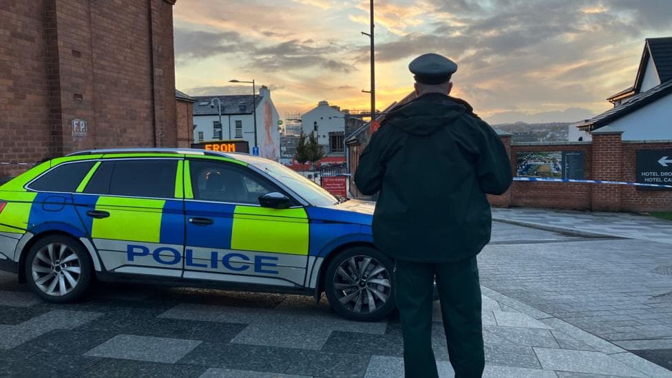A police officer stands with his back to the camera. He is in uniform and standing beside a police car. Police tape can be seen in front of buildings in the background.