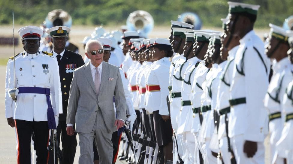 King Charles, wearing a grey suit and sunglasses, arrives at Lynden Pindling International Airport.