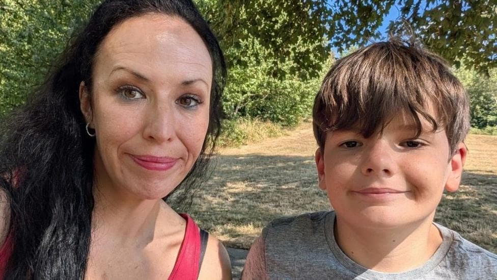 Samantha sits next to her son Oscar. Samantha wears a bright pink vest top and has long dark hair, while Oscar wears a grey T-shirt. Both are facing the camera. Sunlight filters through the leaves overhead, creating patches of light and shadow across the scene. Behind them is a dry, grassy field with green trees and shrubs in the distance under a blue summer sky.