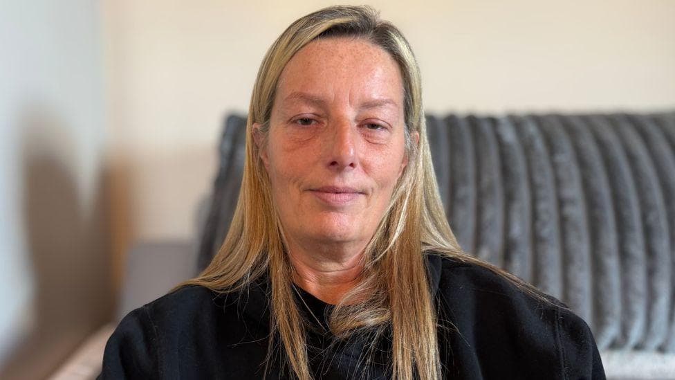 Jill kenning, who has long, blonde hair, wears a black sweater and sits on a grey sofa.