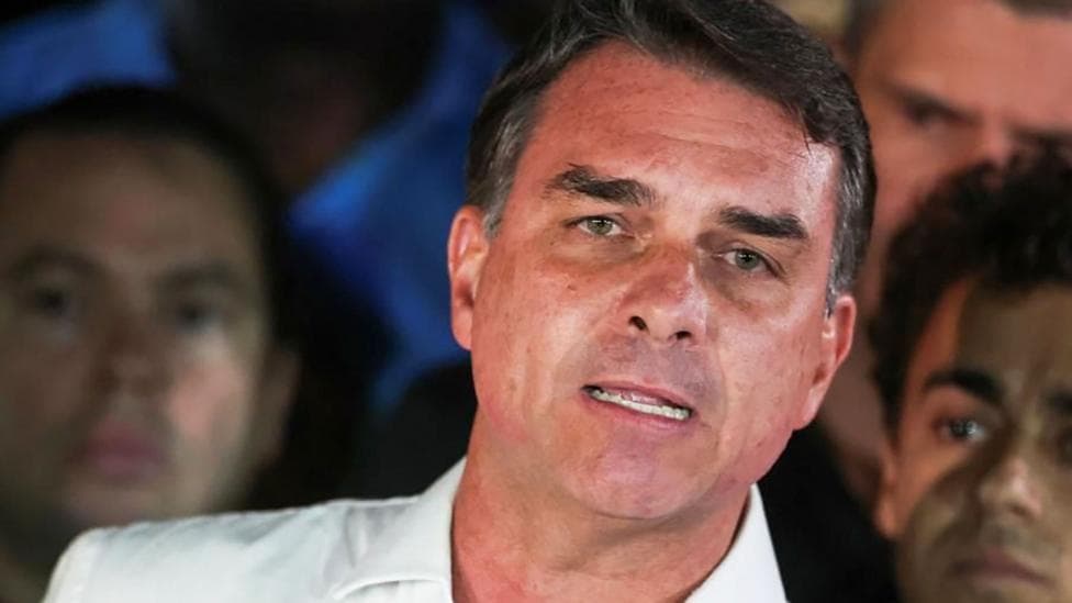 Brazilian presidential candidate Flavio Bolsonaro of the Liberal Party speaks after losing the presidential runoff election, in Brasilia on 25 October, 2026. He is wearing a white shirt with an open collar. He is gesturing with one hand. Behind him, his supporters look on.