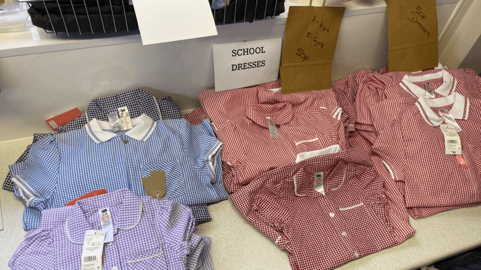 A display of children's school dresses arranged on a shelf in a clothing shop. Several gingham-patterned dresses in red, blue, purple, and navy are folded. A printed sign reading “SCHOOL DRESSES” is positioned at the back of the display, alongside handwritten brown paper labels indicating sizes for different age ranges.