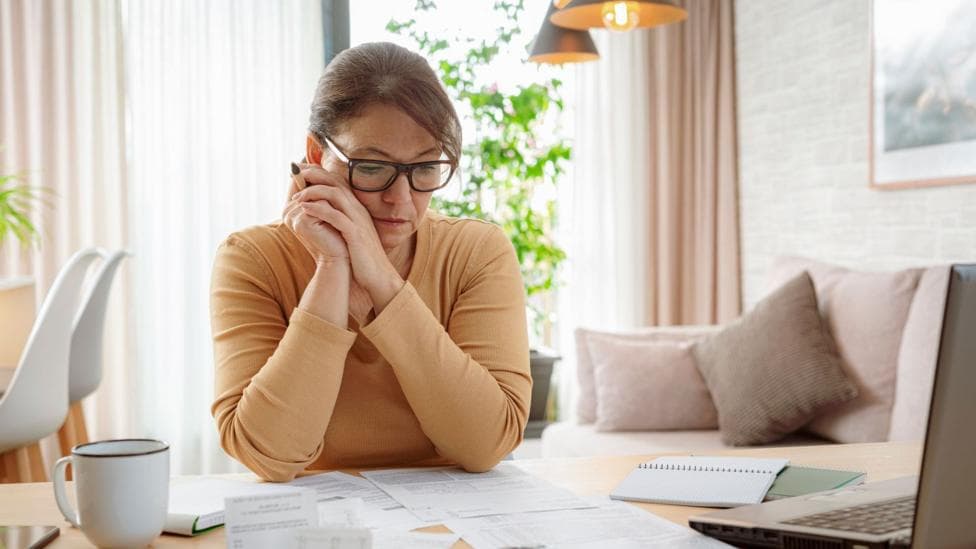 A woman wearing glasses sits at a table covered with paperwork, holding a phone to one side of the face. A laptop, spiral notebook, open folder and white mug are on the table. Behind the table is a sofa with cushions, with a large window with cream curtains, plants and hanging pendant lights.